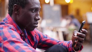 young black man sitting on the street typing on the phone keypad to check messages