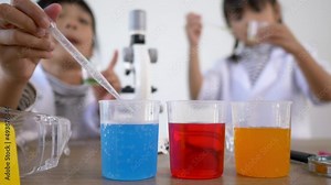 Selective focus at three colors liquids in beakers, blur Asian little girl wearing coat using dropper to suck liquid from glass beaker in background. while studying science chemistry