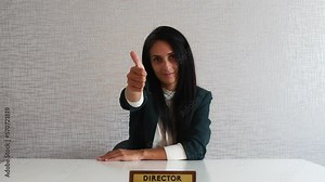 Beautiful adult caucasian woman showing slow motion hand thumbs up hands behind desk, wearing formal office, university clothing, recommending company, sitting on white background