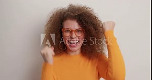 Happy curly haired woman wears orange jumper spectacles makes yes gesture celebrates something feels like winner isolated over white background raises hands up and says yes. Triumph concept.