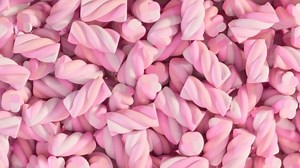 Soft marshmallows falling into a big heap. Lots of pastel pink candy sugar treats in the shape of a swirl or rope lightly falling down. Fluffy confectionery against a pink background.