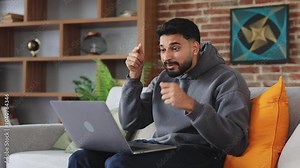 Outraged male in jeans and hoodie pointing with finger at pc screen while shouting with negative expression on apartment background. Tired person throwing aside laptop due to technical malfunction.