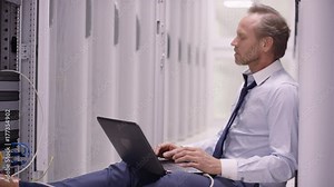 Stressed computer engineer sitting on floor and working on laptop, checking the servers in a data center.
