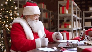 Santa Claus video calling on laptop talking greeting on Merry Christmas, Happy New Year in virtual video online chat meeting on computer sitting at workshop table sending present gift on xmas eve.