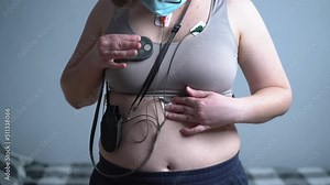 Woman with holter monitor device for daily monitoring of electrocardiogram. Female wearing holter monitor device for daily monitoring of an EKG against the gray wall. Treatment of heart diseases