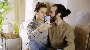 lovely young husband and wife in casual clothes make selfie near pot palm tree and boxes with belongings. Close-up kissing and hugging couple new house owners
