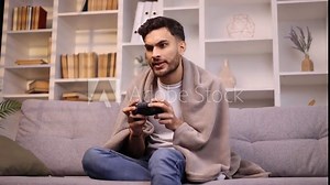 Focused young handsome man with joystick controller playing video games sitting on couch indoors alone Upset male lose the competition at home Defeat Game addiction concept