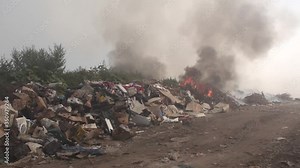 Landfill fire, burning garbage, cardboard boxes, plastic packaging, bags, thick black smoke rising, wider shot of burning, environmental accident, air pollution