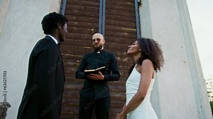 The bride and groom stand in front of the priest on the church threshold