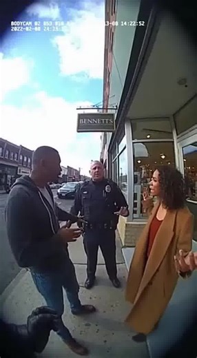 Police body cam footage captures officers in the middle of de-escalating a heated argument on the sidewalk outside a storefront. Voices are raised, hands gesturing, when the sound of an engine suddenly spikes. Tires screech, and in the same moment a car collides with another vehicle in the street, the impact forcing one of them up over the curb. The vehicle slams into the front of the store, glass rattling violently as the facade shudders. Officers instantly shift focus—shouting commands, pushin