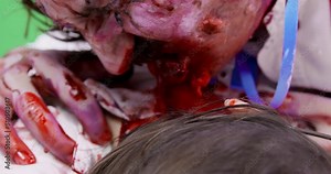 Creepy hungry male zombie with bloody face attacked young woman. He has bitten off pieces of meat from her neck and chewing it, close up. Halloween themed zombie apocalypse party