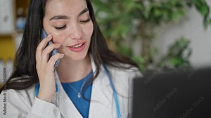 Young beautiful hispanic woman doctor talking on smartphone using computer at the clinic