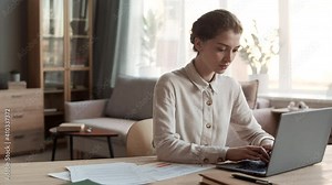 Medium close-up of young professional Caucasian woman wearing formal shirt, sitting by desk in home office, typing on laptop computer, then looking at documents, working from home