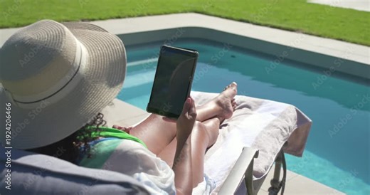 Woman is reclining on lounge chair beside private pool, initiating tablet raise to view screen