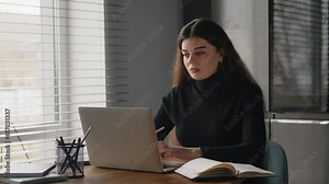 Young business lady sitting at a desk in the office and typing intently on a laptop while writing something in her notebook.