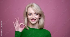A Caucasian woman standing against a vibrant pink background. With her poised demeanor and a captivating smile, she confidently flashes the universally recognized OK gesture.