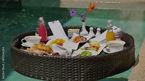 Floating breakfast tray in luxury resort swimming pool with tropical drinks, fresh juice, croissants and flowers. Exotic summer vacation and culinary indulgence.