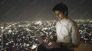 shot of a young Indian male sitting on a terrace of building having a virtual conversation through text message using a laptop against the illuminated cityscape at night