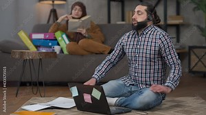 Wide shot portrait of busy serious overworking Middle Eastern man in earphones typing on keyboard analyzing graph sighing and closing laptop. Overworked handsome guy in home office wife at background
