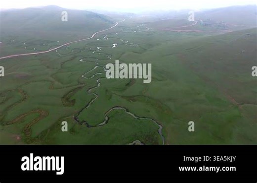 Aerial view of winding meandering river flowing across wide flat green meadow landscape. Curving water bends form natural patterns over expansive grassy plains and open countryside Stock Video Footage - Alamy