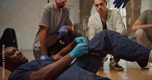 Close-up shooting: A Black male doctor in a blue medical uniform demonstrates the tightening of a medical tourniquet on his leg for public. Practical trainings on first medical aid. Course of a medic