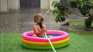 Curious toddler sits in small inflatable pool under tropical downpour, holding hand sprinkler. Enjoying warmth of heavy rain, he playfully experiments with water, sprinkling it around.