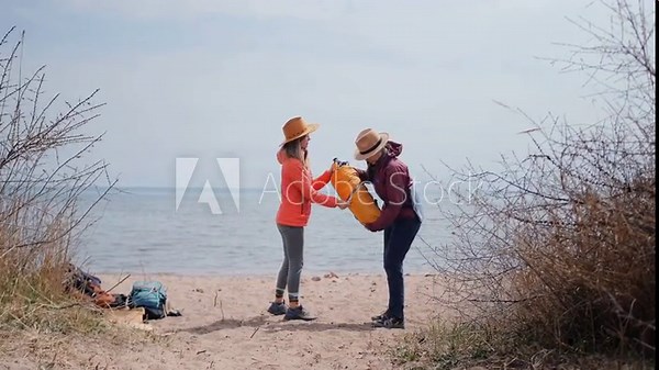 two women standing front view beach unpacking tent from backpack near lake, preparing campsite together, outdoor travel activity, hiking trip, teamwork, camping setup and exploration