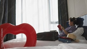 Asian woman in bathrobe and hair curlers sitting on cozy bed, reading travel guide book, prepare clothes into luggage, Making check list of things to pack for travel, light through curtain the window.
