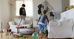 Three kids are energetically jumping on a white couch surrounded by toys and household items in a disordered living room, with sunlight streaming through the window.