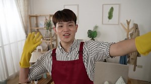 asian male vlogger wearing apron and gloves waving hand at mobile cam and talking while vlogging about his housecleaning routine in a bright living room at home