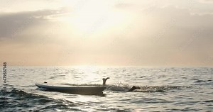 Wave makes sitting young woman wearing bikini swimsuit to fall from stand-up paddle board into sea water against sky with bright sunlight