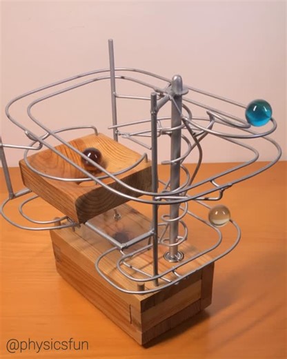 physicsfun on Instagram: "Rolling Marble Sculpture: marbles perform gymnastics as potential energy of gravity is converted to kinetic energy. An elegantly designed auger conveyor, an Archimedes’ Screw, brings the marbles back to the top (powered by an AA battery and a small motor). Conservation of angular momentum produces a simulation of decaying orbits as the marbles whirl down the the main funnel shaped feature. Made with recycled wood by artist Bogdan Krupa ➡️ Follow the link in my profile f