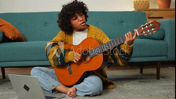 African American woman learning guitar watching tutorial on laptop at home. Black girl playing guitar singing song learning online music lessons. Artistic woman playing acoustic guitar learning chords