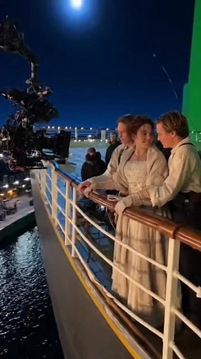 A rare behind-the-scenes moment from a period-film dance sequence. The actors rehearse their lively dance steps in a rustic wooden hall set, surrounded by glowing lanterns, wooden beams, and warm cinematic lighting. Crew members work just inches away — the boom operator holds the microphone above the actors while the camera team tracks the movement with a large cinema rig aimed directly at the center of the dance floor. Background performers clap and smile, creating a cheerful, energetic atmosph