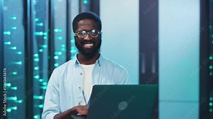 Nice-looking african IT specialist using a laptop computer working in database server room smiling with joy at camera. Modern people. Data center.
