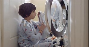 A young boy in patterned pajamas is carefully placing clothes into an open front-loading washing machine, demonstrating independence and responsibility.