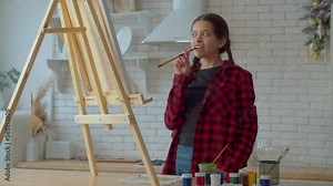 Thoughtful creative lovely Hispanic teenage girl painter with paintbrush in mouth standing near easel, searching for artistic inspiration and thinking about ideas for painting at home.