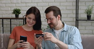 Head shot young man in glasses dictating credit banking card payment information to focused millennial woman, entering cvv code in mobile shopping app, happy couple involved in purchasing goods online