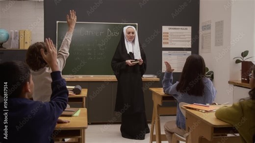 Front view shot of smiling young woman as Catholic nun dressed in traditional habit holding bible and giving speech on career day in school classroom with group of children raising hands in foreground
