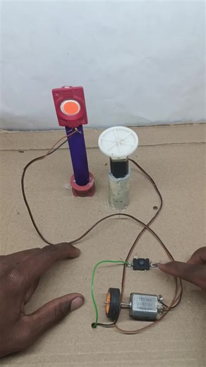 It likely contains a simple push-button switch wired in series with the motor and a battery pack (not visible), where the LED illuminates to confirm power is flowing. The orange glow provides visual feedback during operation, common in homemade science projects for safety and ease. Wires from the box connect directly to the motor and ground, enabling the electrostatic charging process.