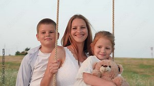 Family of mother and little children playing swings portrait in summer field at sunset. Smiling mother with children swings in rural meadow. Happy mother with girl and boy on swings country park