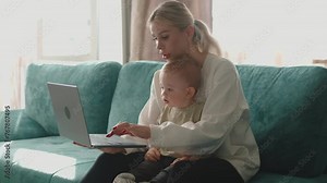 Young smiling mom sitting on sofa in front of the computer laptop screen, working remotely on laptop, writing messages, holding her little son on lap, child is polite