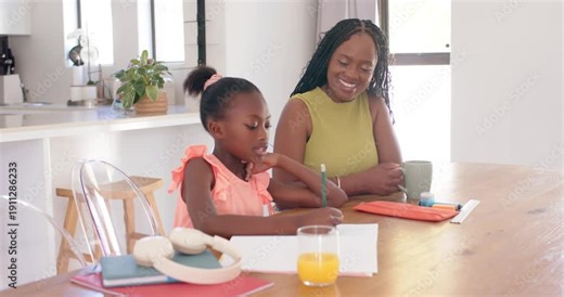 African American mom and daughter sitting at home, child doing worksheet, mom smiling with mug