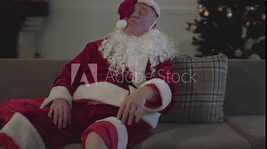 Drunk old man with false white beard in the costume of Santa Claus lying on the sofa at home, waving hand. Bad Santa. Alcoholism, depression, negativism, loneliness
