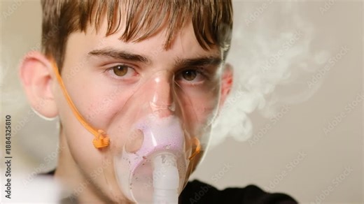 Caucasian teenage boy looks at camera and breathes nebulized medical mixture from inhaler using silicone face mask. Calm and questioning patient look. Treatment of lung diseases using inhaler.