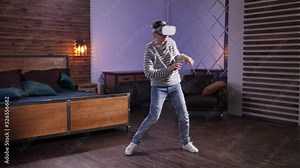 Cute teenage boy using augmented-reality headset while dancing in popular funny style at home. Advanced teenager in hoodie and jeans having fun in virtual reality during domestic leisure