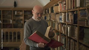 Mature professor is reading book standing in antique library, bearded man wearing eyeglasses is holding red exhibit, looking, having time on background of bookshelves in vintage room. Concept