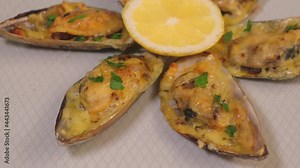 Baked Greenshell mussels in half a shell with cheese, parsley and lemon on a white plate. Traditional hot appetizer. Close-up, top view, rotation.