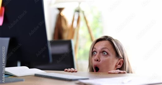 Woman hides behind an office desk and looks at computer monitor in shock. Bad news emotions of fear and horror on internet