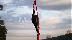 Silhouette of Female circus artist showing her flexibility and splits with red aerial silk on the dramatic sky background in slow motion. Concept of healthy lifestyle